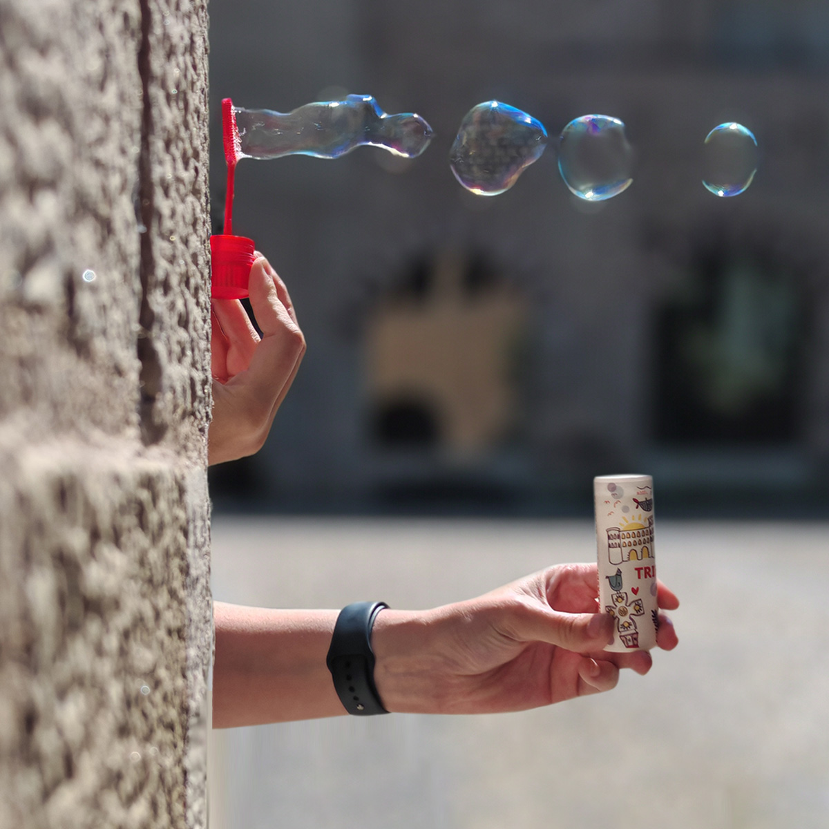 Fläschchen mit Seifenblasenflüssigkeit Zwei Hände, die vor einer Wand hervorschauen,  halten ein Fläschchen mit Seifenblasenflüssigkeit und machen Seifenblasen